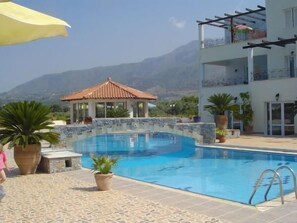 A swimming pool with a tiled deck, a covered gazebo, and a building with balconies. - Pinelopi Apartments (Apokoronas)