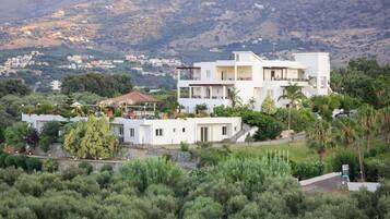 A multi-story residential building with balconies, surrounded by greenery and a hilly landscape.