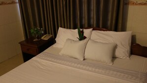 A neatly made bed with white linens and pillows, a bedside table with a telephone and a small plant, and a curtain in the background. - Relax Guesthouse (Phnom Penh)
