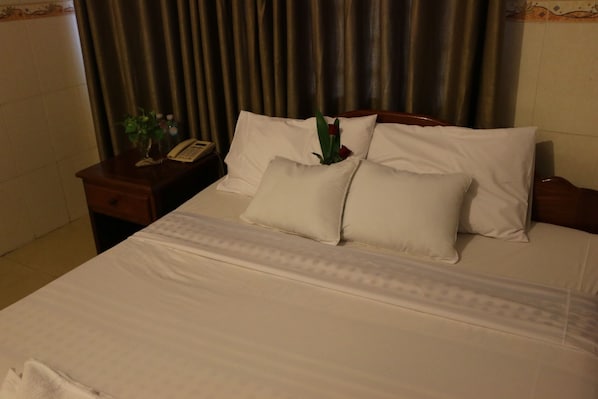 A neatly made bed with white linens and pillows, a bedside table with a telephone and a small plant, and a curtain in the background.