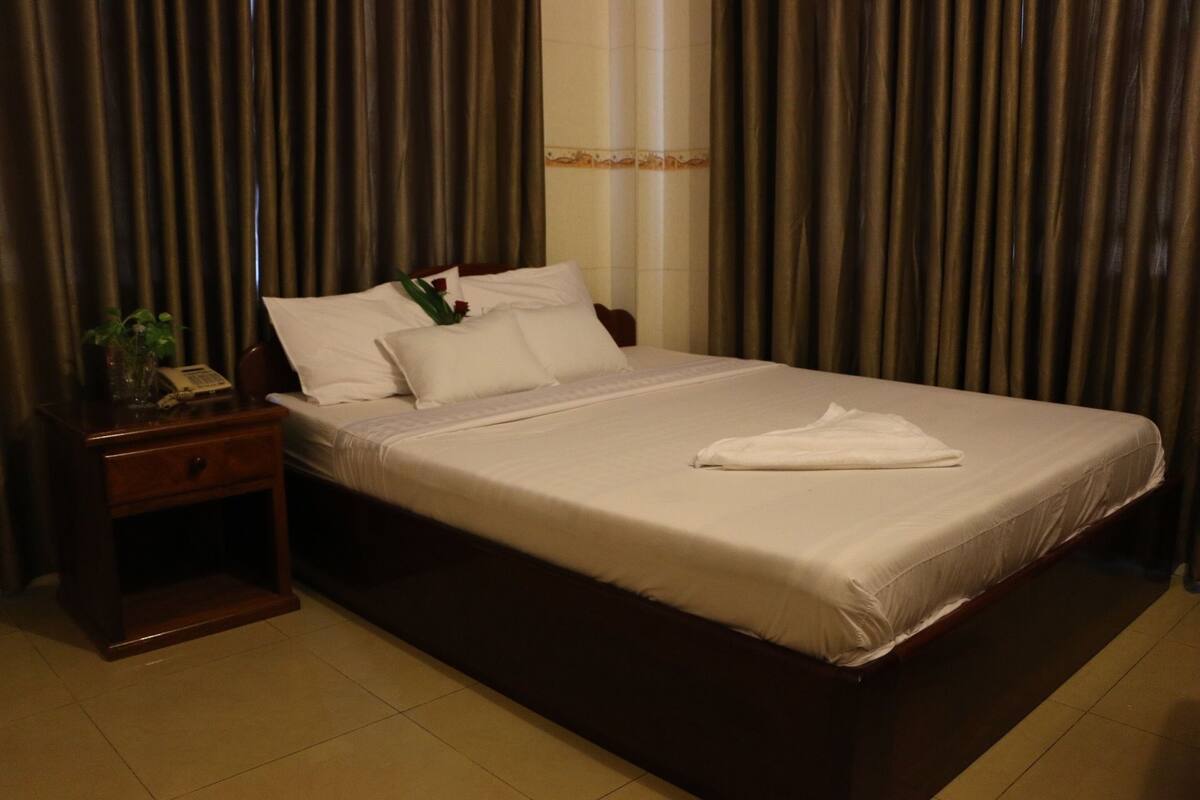 A neatly made bed with white linens, a wooden headboard, and a bedside table with a telephone and a vase with flowers.