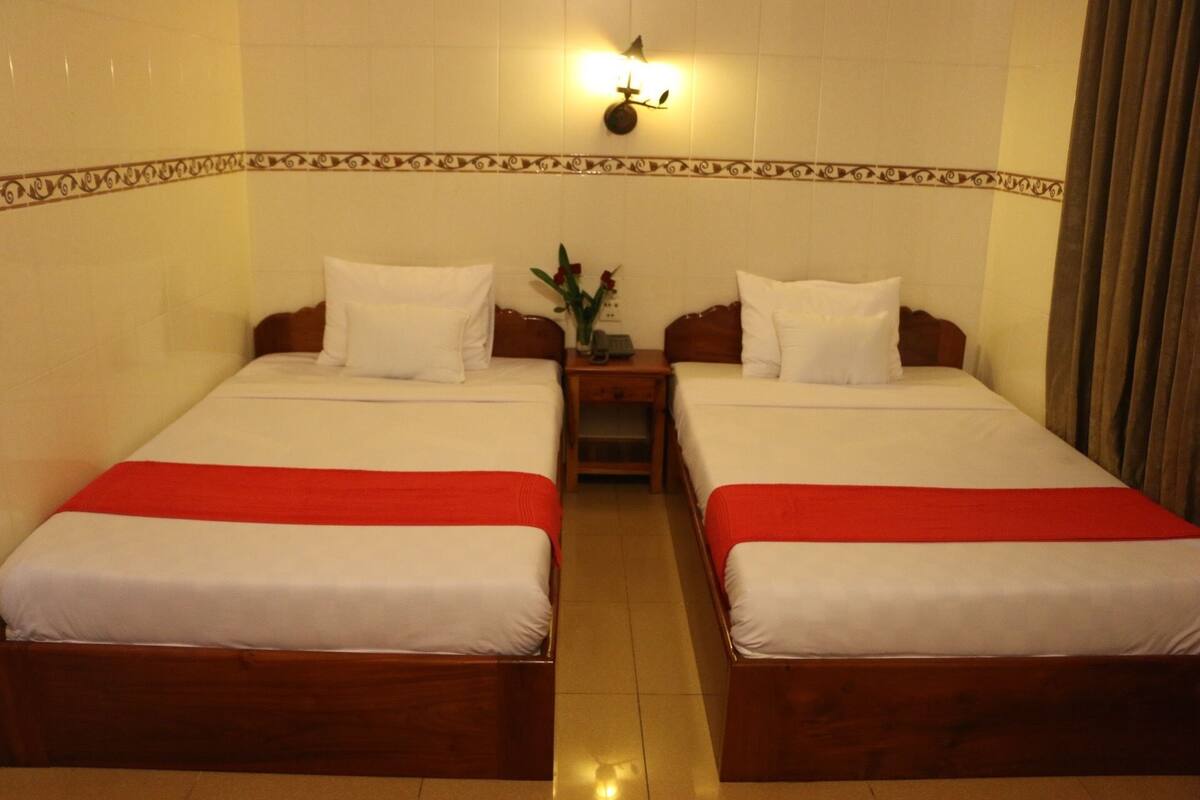A hotel room with two single beds, each with white bedding and a red blanket. There is a nightstand between the beds with a vase of flowers and a lamp. The room has a tiled floor and a decorative border on the wall.