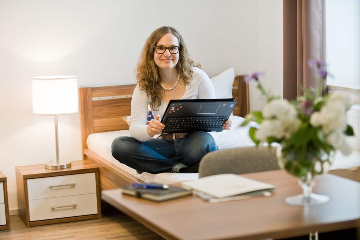 A person sitting on a bed with a laptop, a bedside table with a lamp, and a vase with flowers.