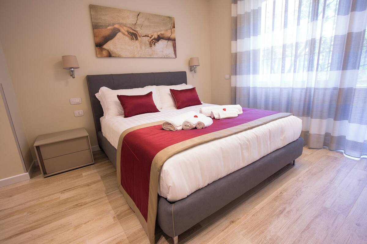 A neatly made bed with red and white linens, a bedside table, and a painting on the wall.