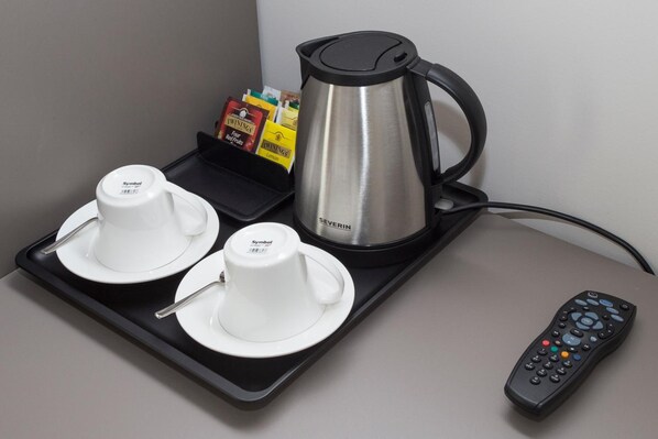 Un plateau avec deux tasses et sous-tasses blanches, une bouilloire électrique argentée, une télécommande et un présentoir de sachets de thé.