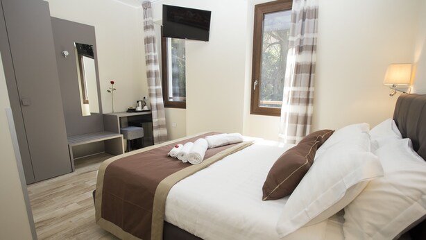 Quarto de hotel bem arrumado, com uma cama, televisão presa na parede, escrivaninha e janela com cortinas.