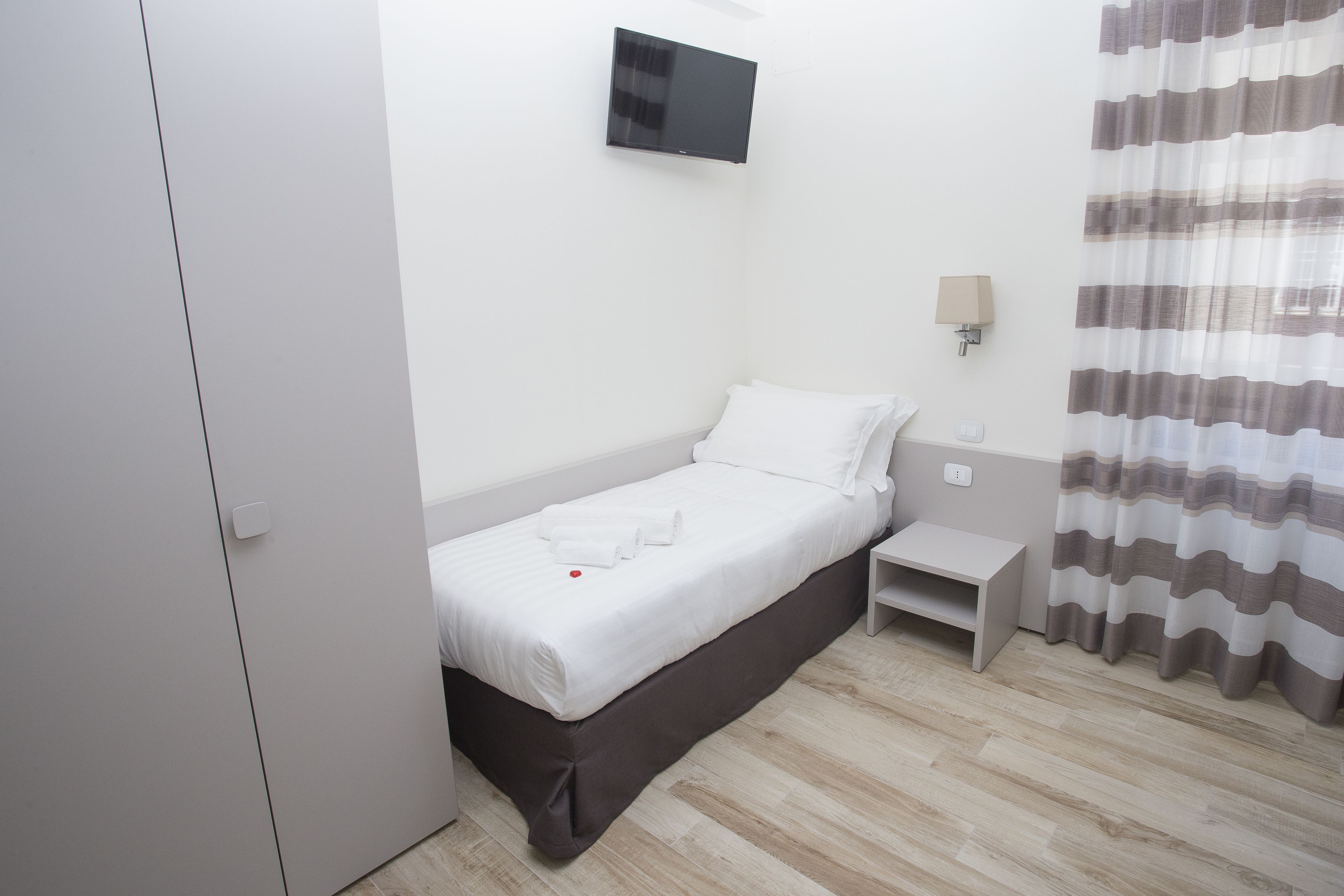A hotel room with a single bed, a bedside table, a TV mounted on the wall, and a curtain.