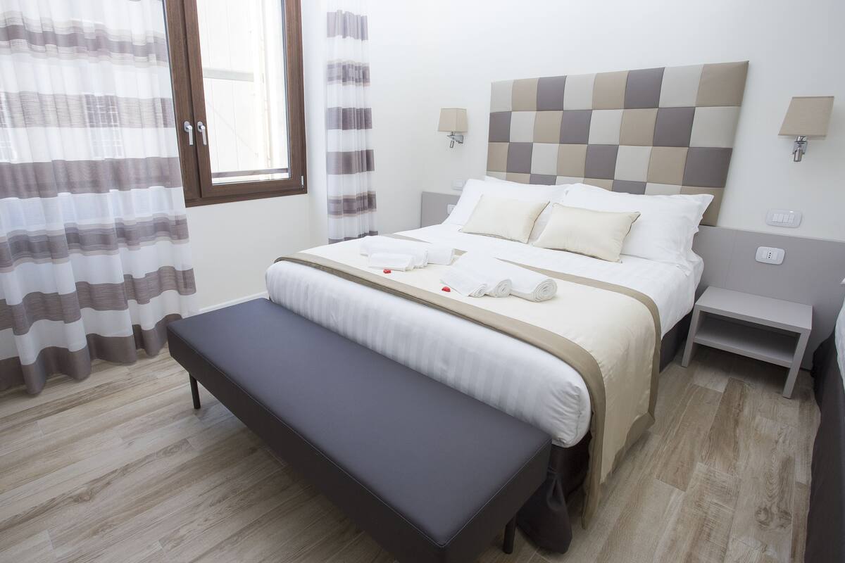 A neatly arranged hotel room with a bed, bedside tables, a bench, and a window with curtains.