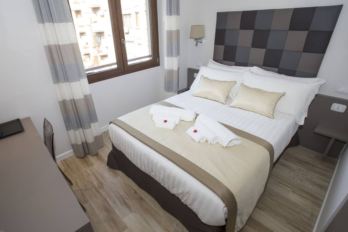 A neatly made bed with white and beige linens, a bedside table with a lamp, and a window with curtains.