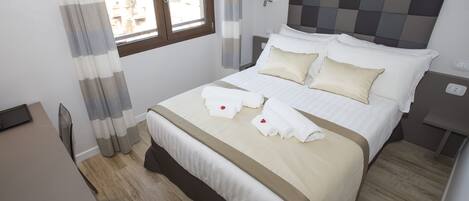 Una cama bien hecha con ropa de cama blanca y beige, una mesita de noche con lámpara y una ventana con cortinas.