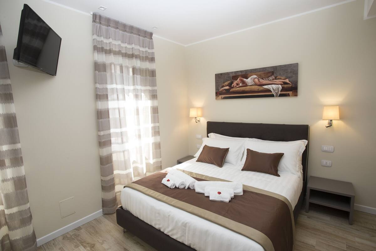 A neatly made bed with a brown headboard, two bedside lamps, and a painting above it.