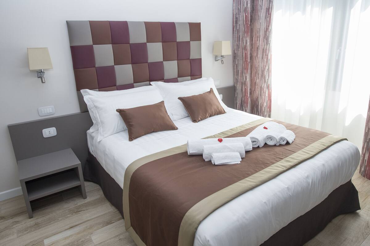 A neatly made bed with a brown headboard, white pillows, and a brown bedspread. There are two bedside tables, one with a lamp and the other with a power outlet.