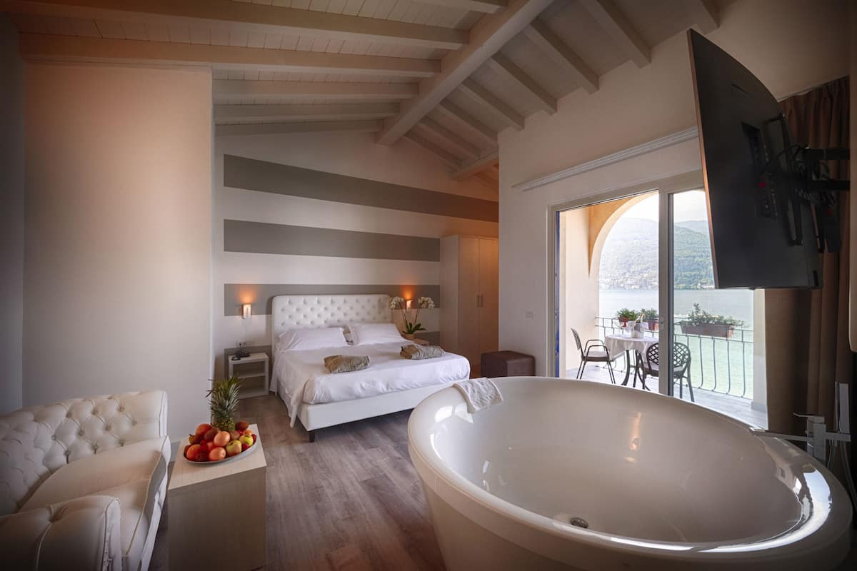 A bedroom with a large bed, a sitting area with a sofa, a bathtub, and a balcony with a view of the water.