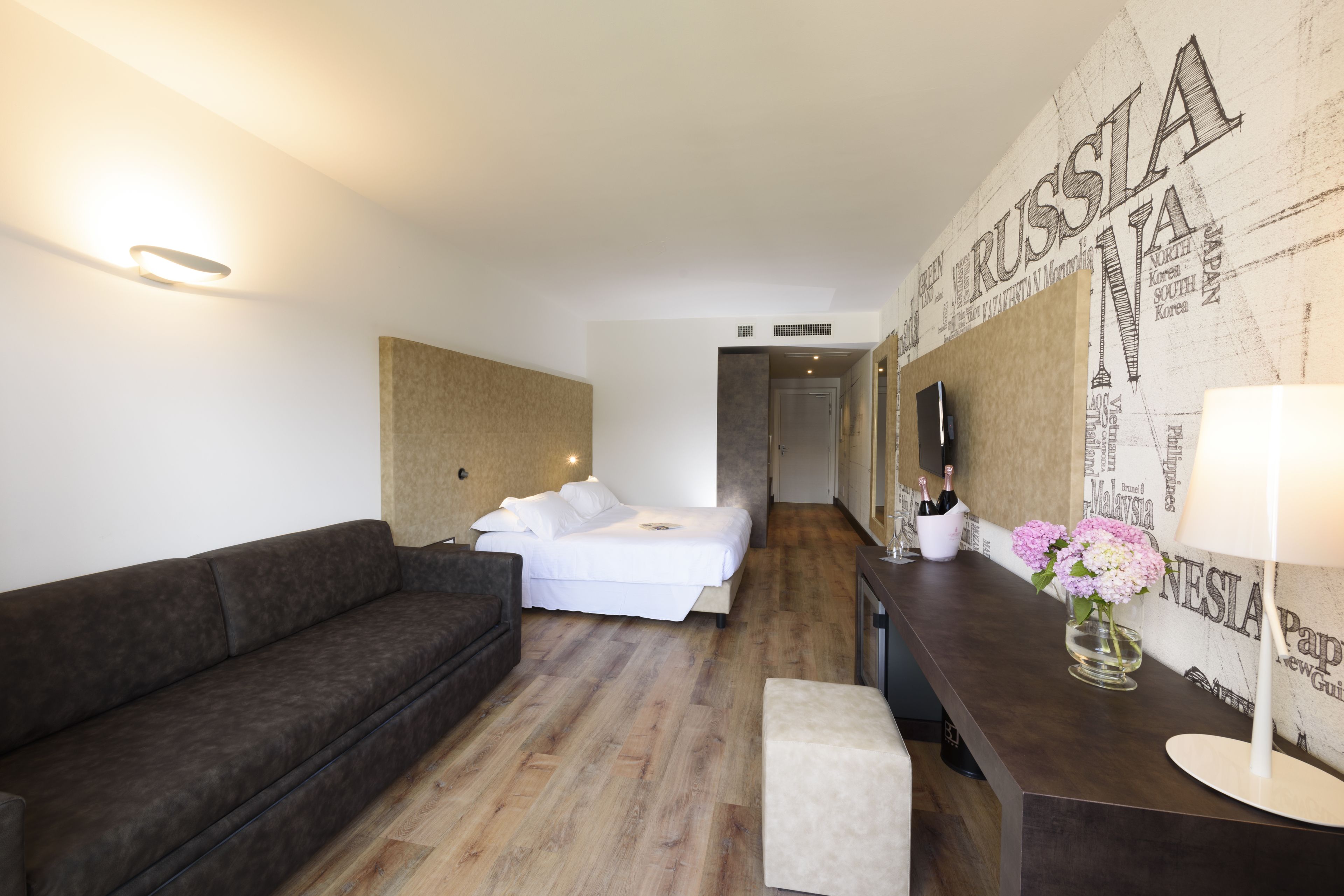 Une chambre d’hôtel moderne avec un plancher en bois, un canapé, un lit, un bureau avec une lampe et des fleurs, et un mur orné d’une carte de la Russie.