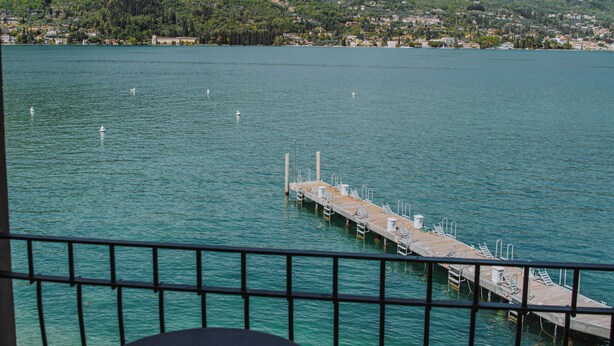 Un balcón con mesa y sillas que da a un lago con un muelle y montañas al fondo.