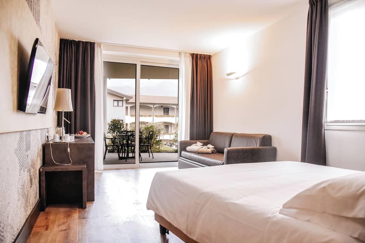 A hotel room with a bed, a desk with a lamp, a chair, and a view of the outdoor area.