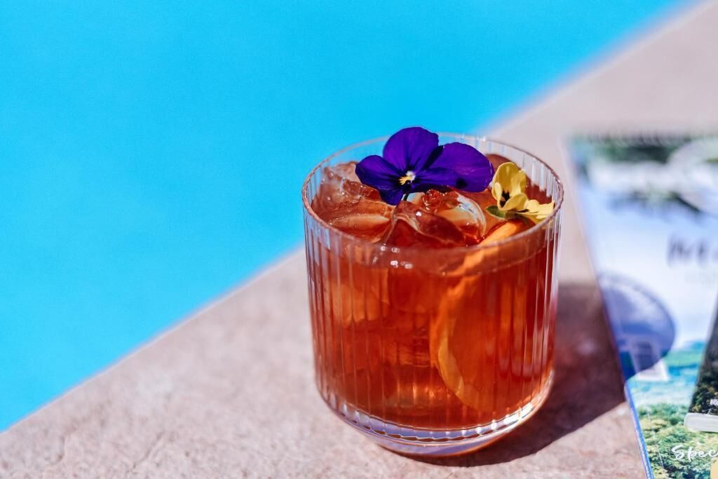 A glass of amber-colored beverage with a purple flower garnish, placed on a surface near a pool.