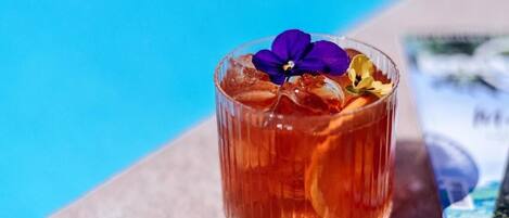 A glass of amber-colored beverage with a purple flower garnish, placed on a surface near a pool.