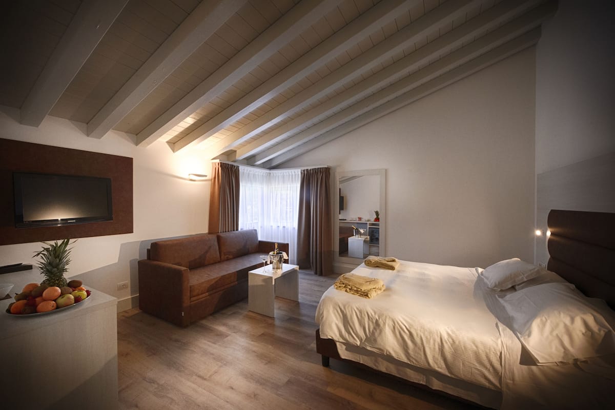 A modern hotel room with a wooden ceiling, a bed, a sofa, a small table with fruit, and a TV mounted on the wall.
