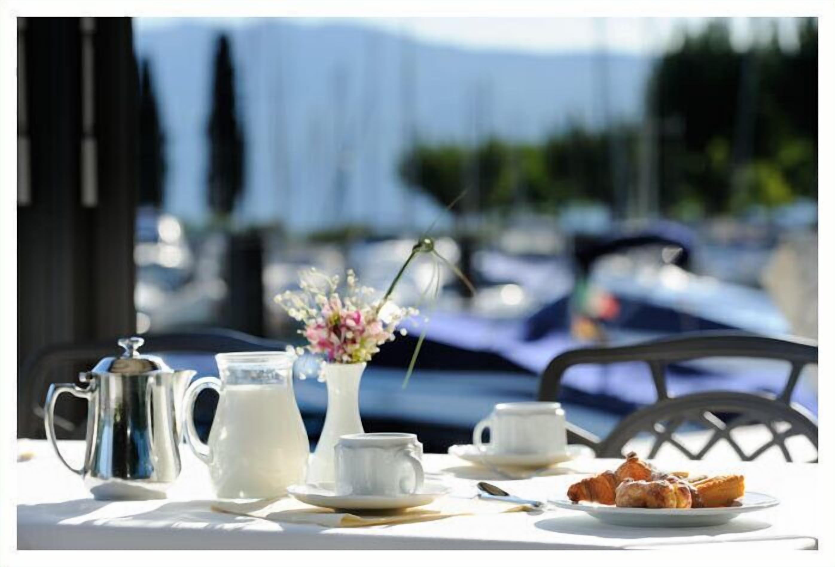 A table set with a milk jug, a pitcher, a vase with flowers, cups, and a plate of pastries, with a marina view in the background.