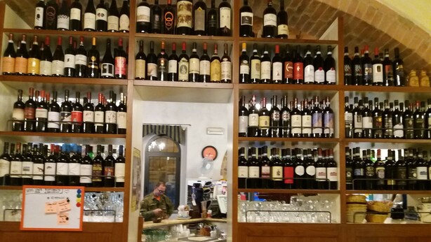 A wine cellar with shelves of wine bottles and a person in the background. - Oasi del grillo (Castelnuovo Berardenga)