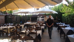 An outdoor dining area with tables and chairs under large umbrellas, a grill, and a brick wall in the background. - Oasi del grillo (Castelnuovo Berardenga)