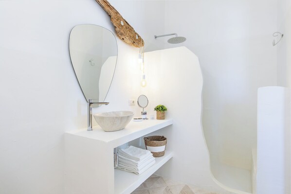 Un baño moderno con un espejo grande, un lavabo, una ducha y un tocador blanco con toallas y una planta pequeña.