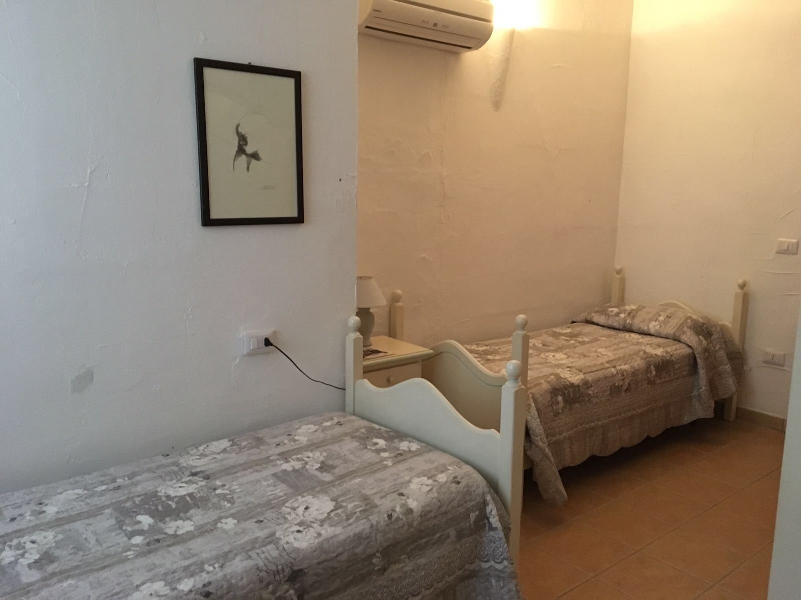 Un dormitorio pequeño con dos camas individuales, una mesita de noche con lámpara y un equipo de aire acondicionado instalado en la pared.