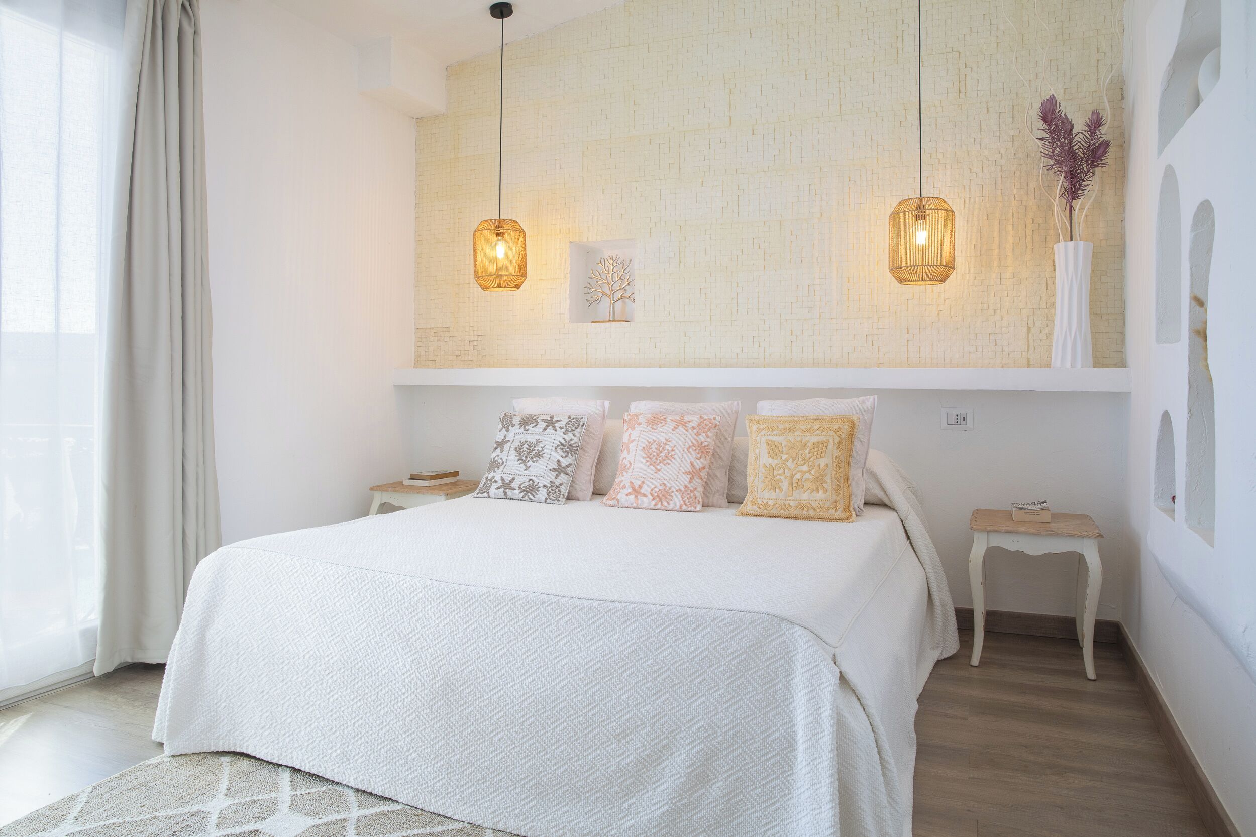 A bedroom with a large bed, two bedside tables, a nightstand, a wall-mounted shelf, and two hanging pendant lights.