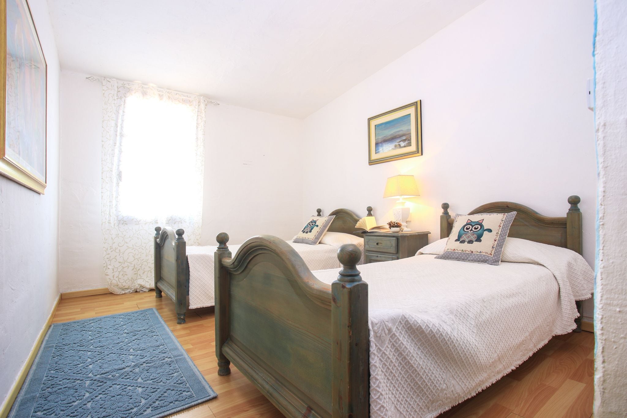 Une chambre à coucher avec deux lits en bois, une table de chevet avec une lampe, un tableau encadré au mur et une fenêtre avec des rideaux.