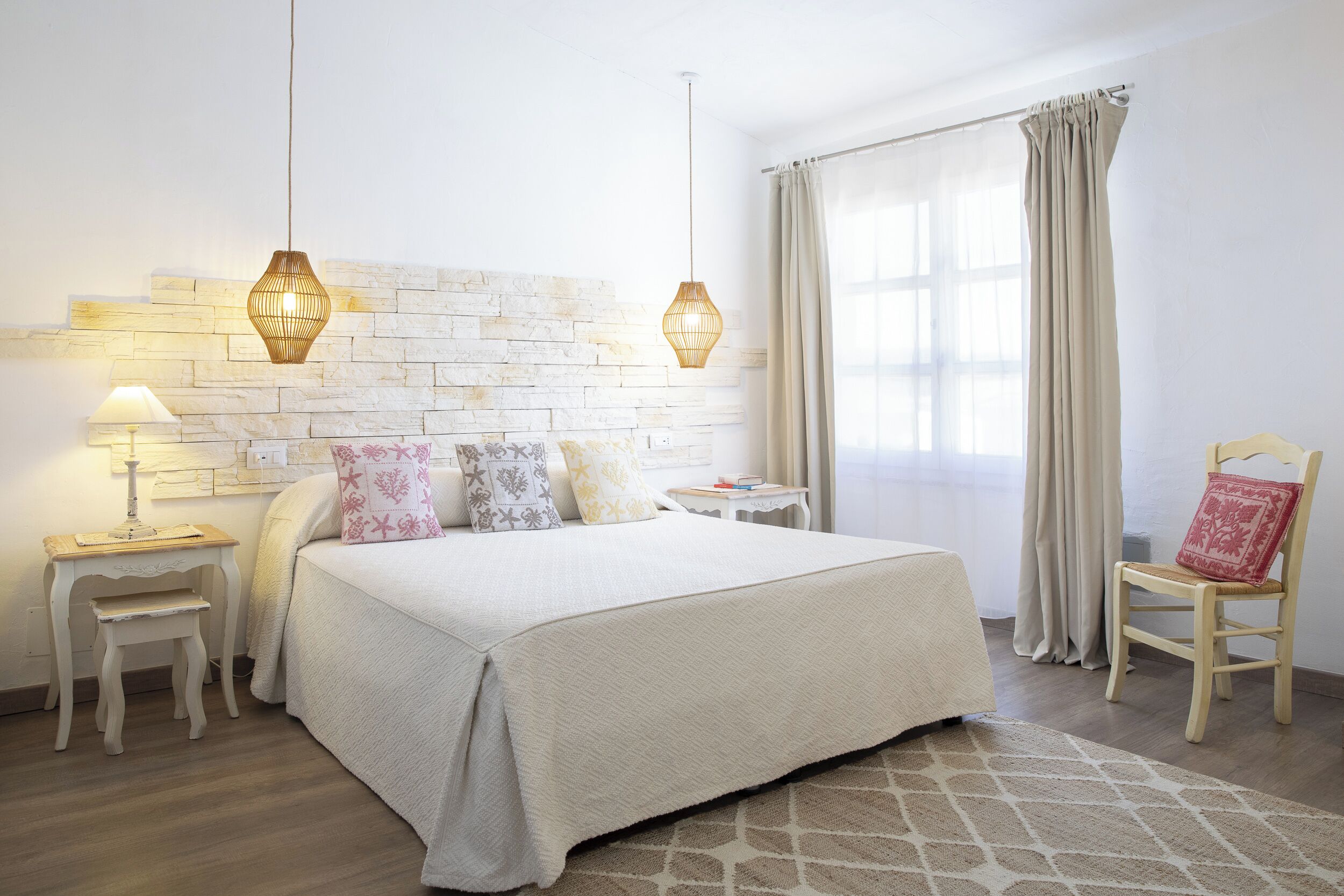Quarto bem arrumado com uma cama grande, duas mesas de cabeceira, um banquinho pequeno, uma cadeira e uma janela com cortinas.