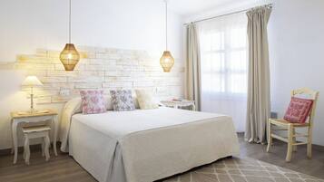 Quarto bem arrumado com uma cama grande, duas mesas de cabeceira, um banquinho pequeno, uma cadeira e uma janela com cortinas.