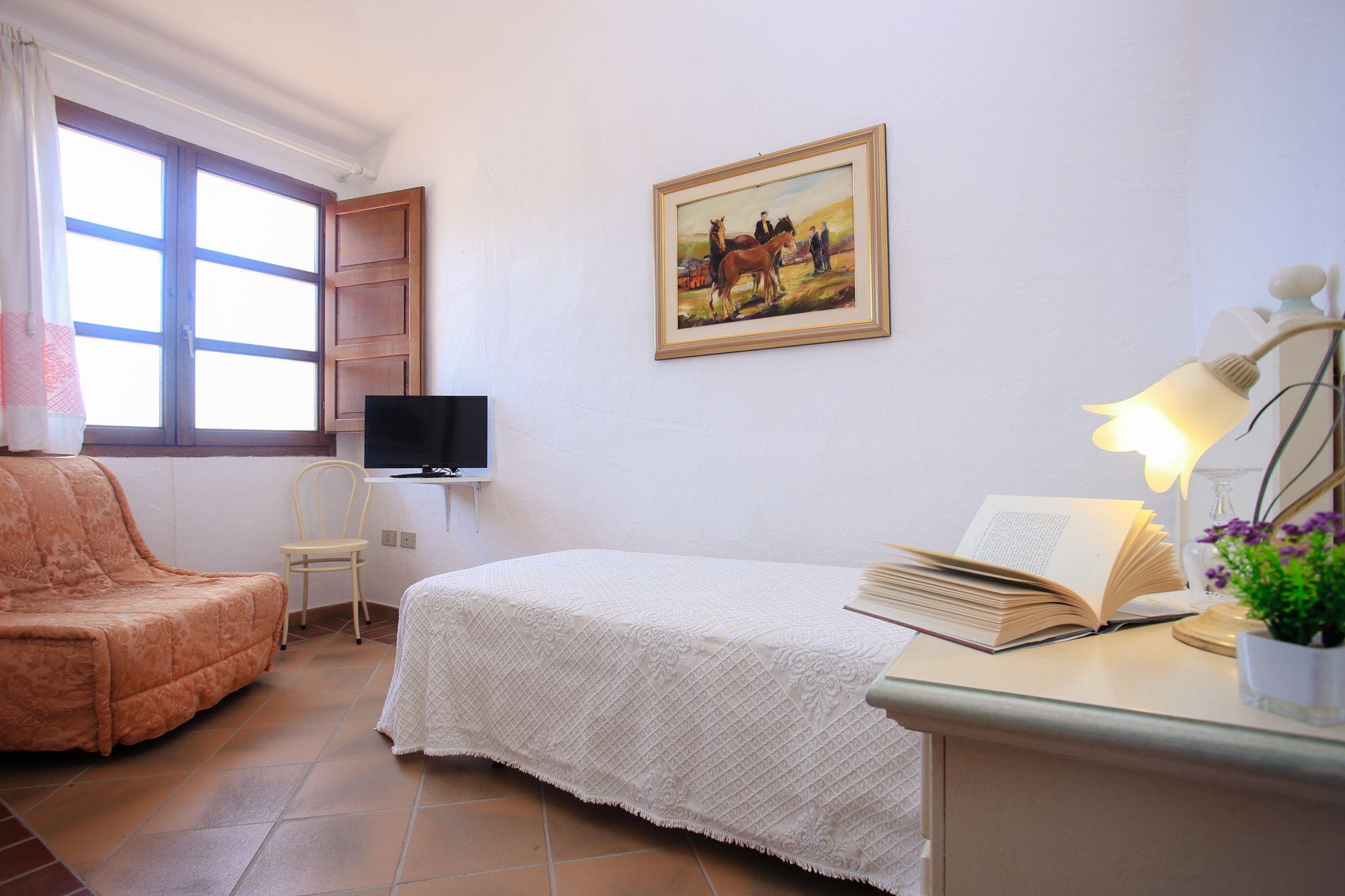 Un dormitorio con cama, una silla, un escritorio con lámpara, un televisor y un cuadro en la pared.