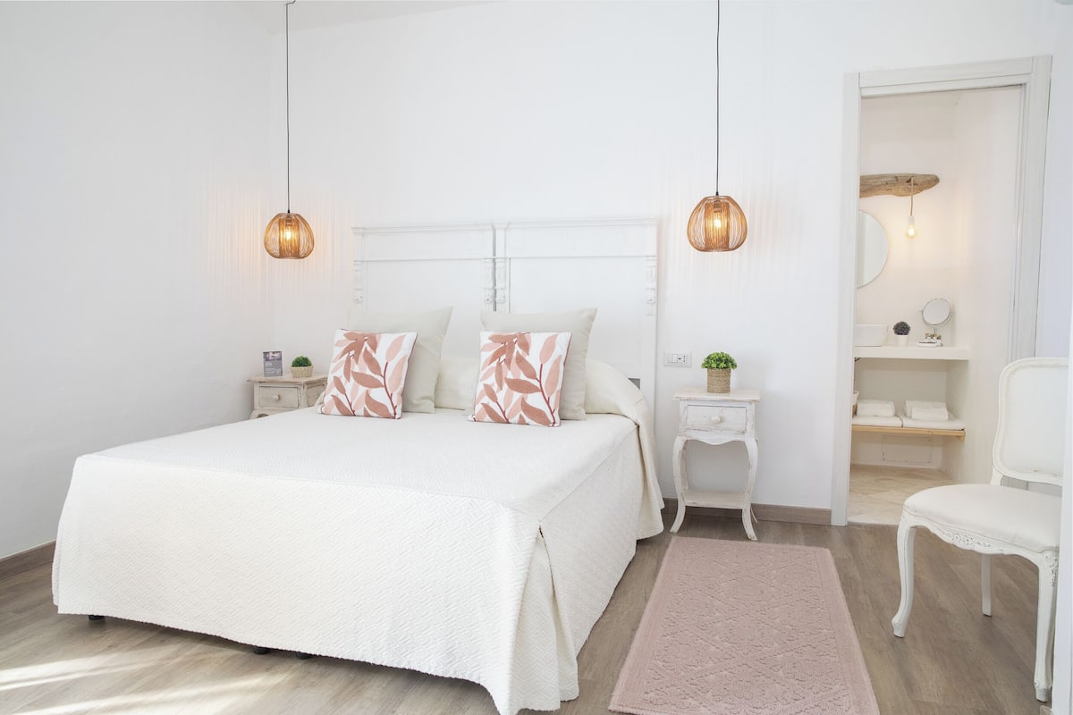 A bedroom with a large bed, two bedside tables, a chair, a small table with a plant, and a mirror above the door.