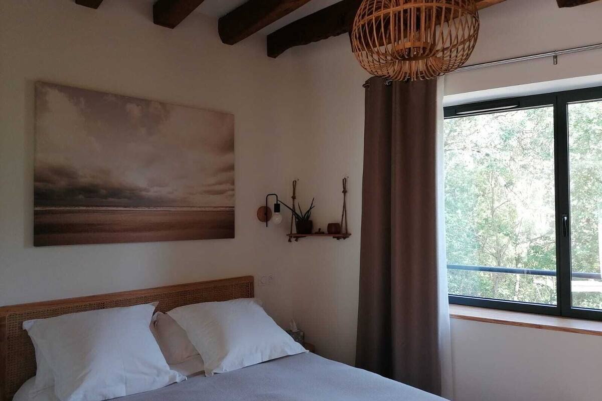 A bedroom with a bed, a painting, a wall-mounted light fixture, and a window with curtains.