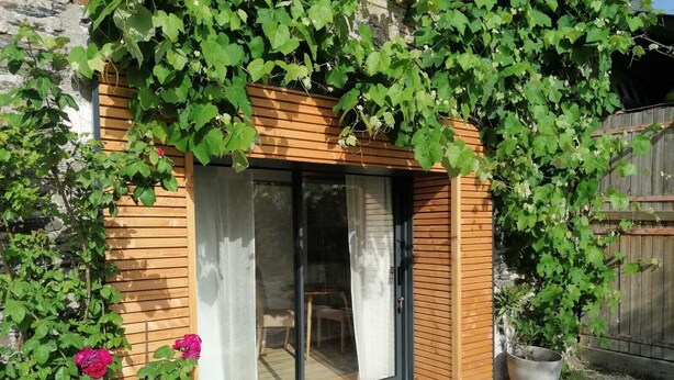 A modern house with a wooden exterior, a sliding glass door, and a garden area with potted plants and flowers. - La Petite Boire - chambres d'hôtes (Murs-Erigne)