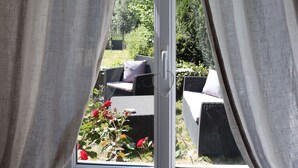 A room with a large window, curtains, and a view of outdoor furniture and greenery. - La Petite Boire - chambres d'hôtes (Murs-Erigne)