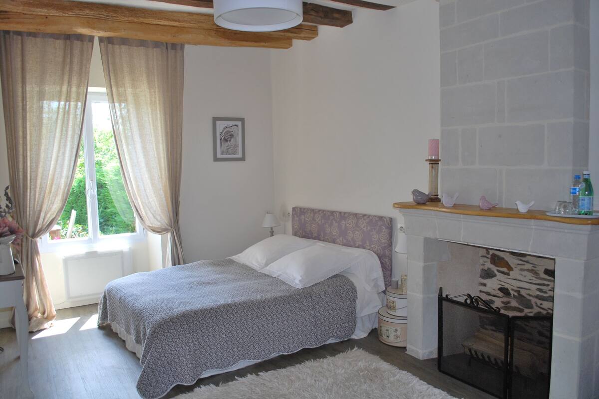 A bedroom with a bed, a fireplace, a window with curtains, and a framed picture on the wall.