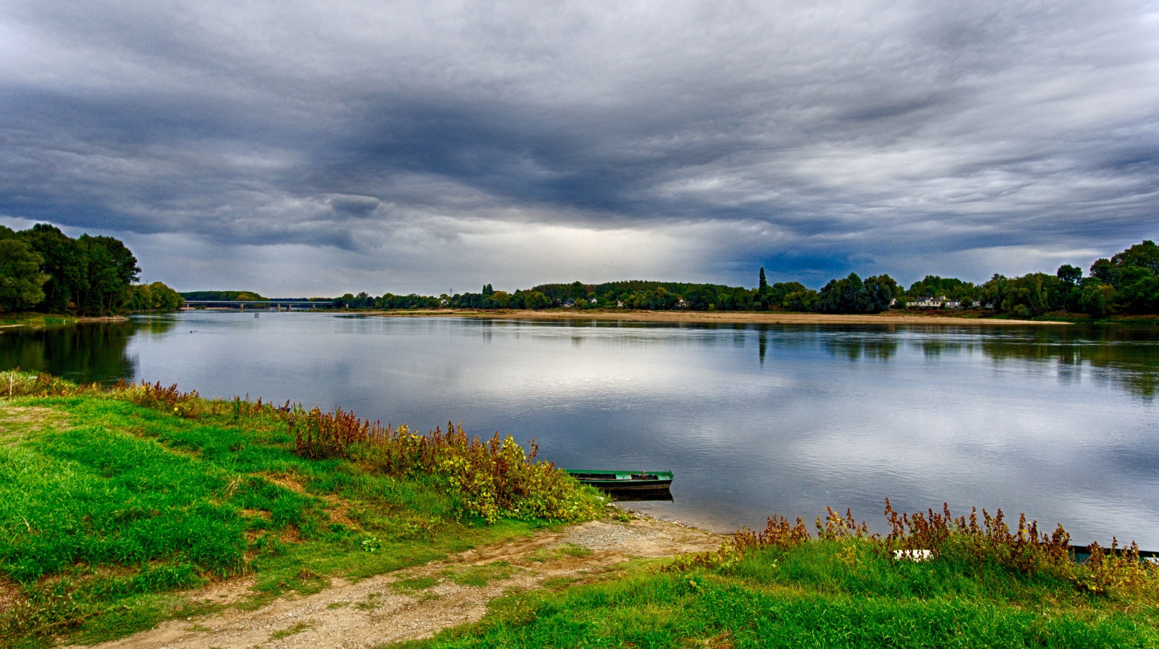 Ein ruhiger Fluss mit einem am Ufer vertäuten Boot, umgeben von Grün und einem bewölkten Himmel.