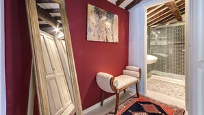 A hallway with a red wall, a large ornate mirror, a bench, and a glass door leading to a bathroom. - LM Suite Spagna (Rome)