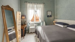 A bedroom with a bed, a chair, a desk with a lamp, a mirror, and a window with curtains. - LM Suite Spagna (Rome)