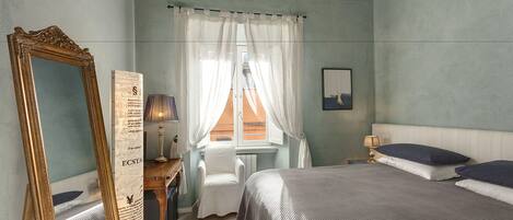 Un dormitorio con cama, una silla, un escritorio con lámpara, un espejo y una ventana con cortinas.