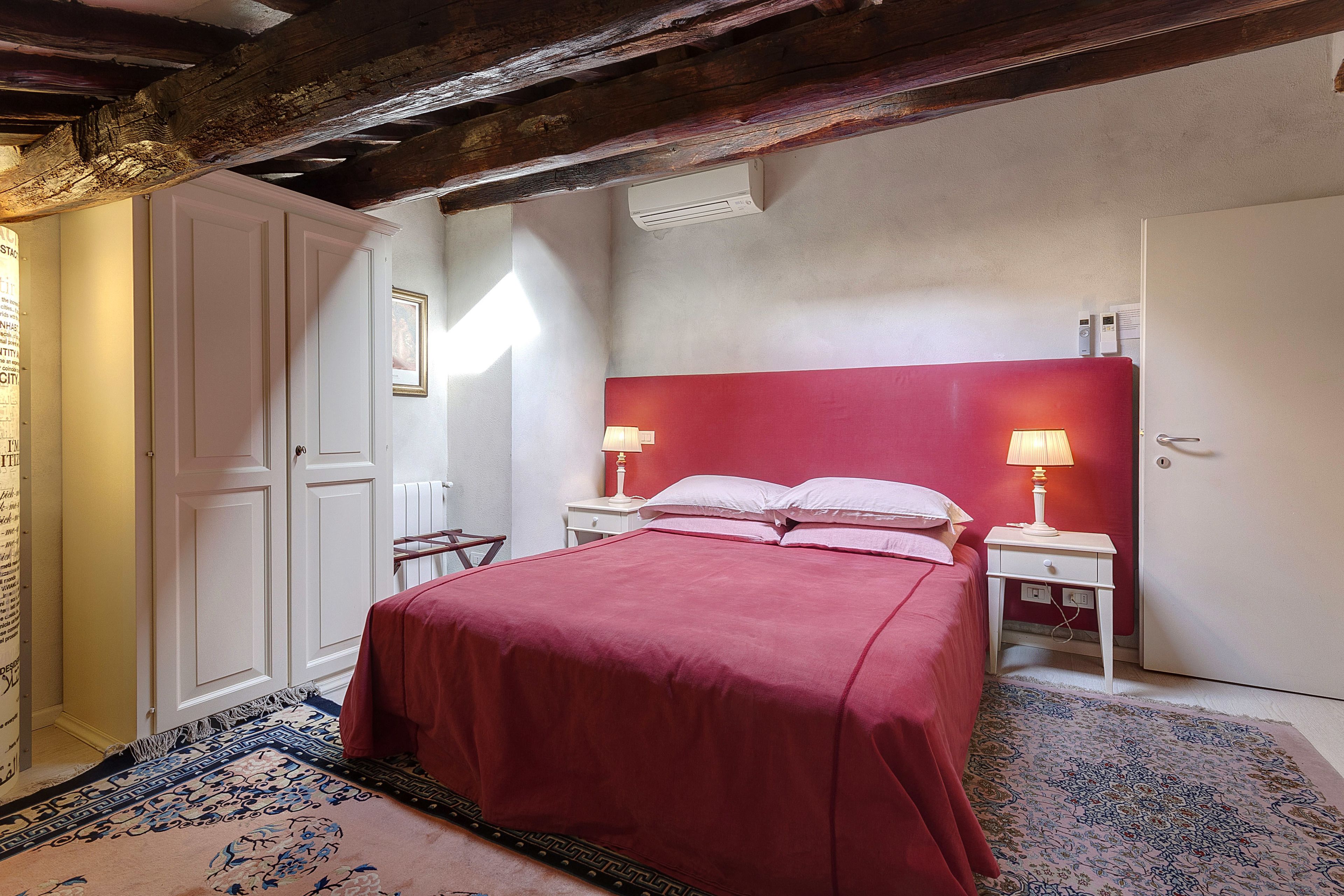 A bedroom with a red bedspread, white walls, a wooden ceiling, and a patterned rug.