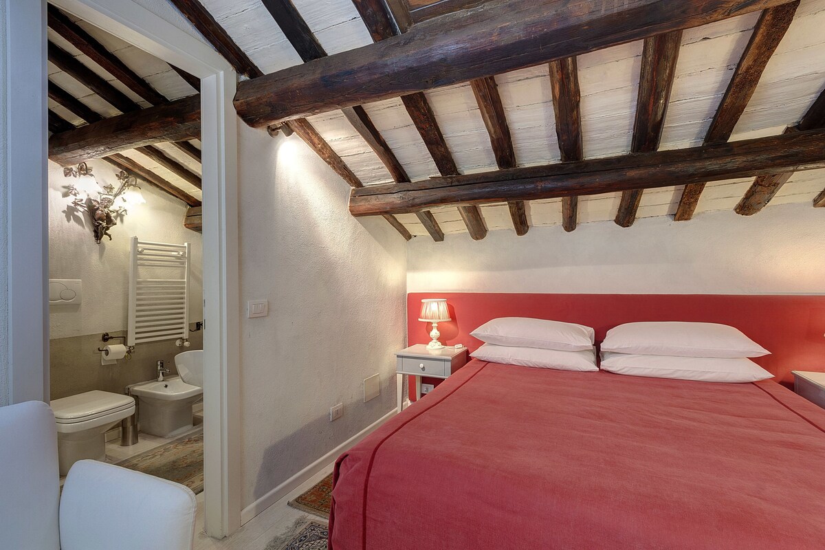 A bedroom with a red bedspread, a nightstand with a lamp, a white bedside table, and a wooden ceiling.