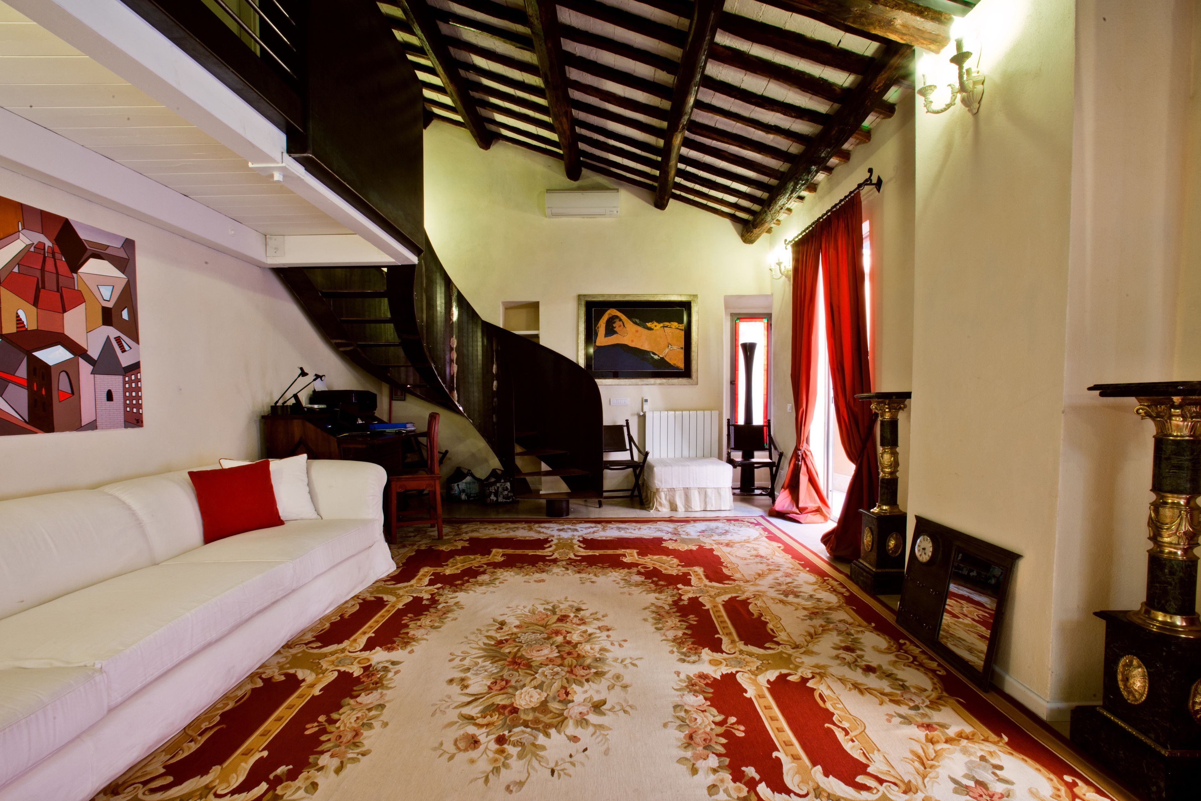 A spacious living room with a large red and beige carpet, a white sofa with red cushions, a staircase leading to an upper floor, and a chandelier.