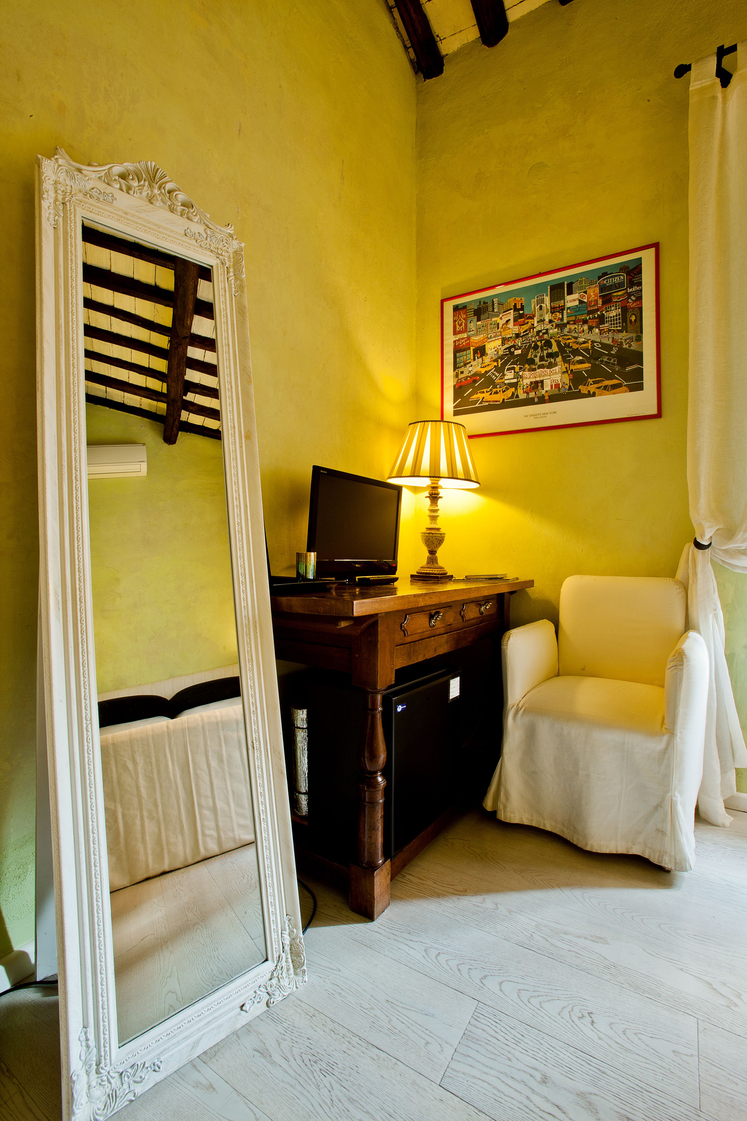 Ein Zimmer mit Spiegel, Schreibtisch mit Lampe, Stuhl und einem gerahmten Bild an der Wand.