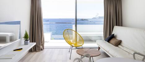 A modern living room with a white sofa, a yellow chair, a small round table, and a TV on a white stand. The room has a large sliding glass door leading to a balcony with a view of the sea.