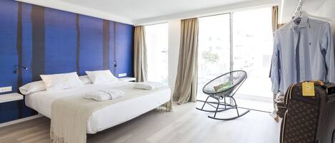 Una habitación de hotel moderna con una cama grande, un sillón mecedora, una maleta y vistas de la ciudad a través de amplias ventanas.
