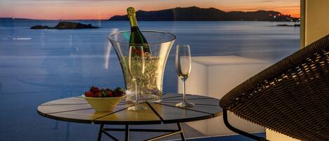 A table with a bottle of champagne, a bowl of strawberries, and a glass on a balcony overlooking a body of water at sunset.