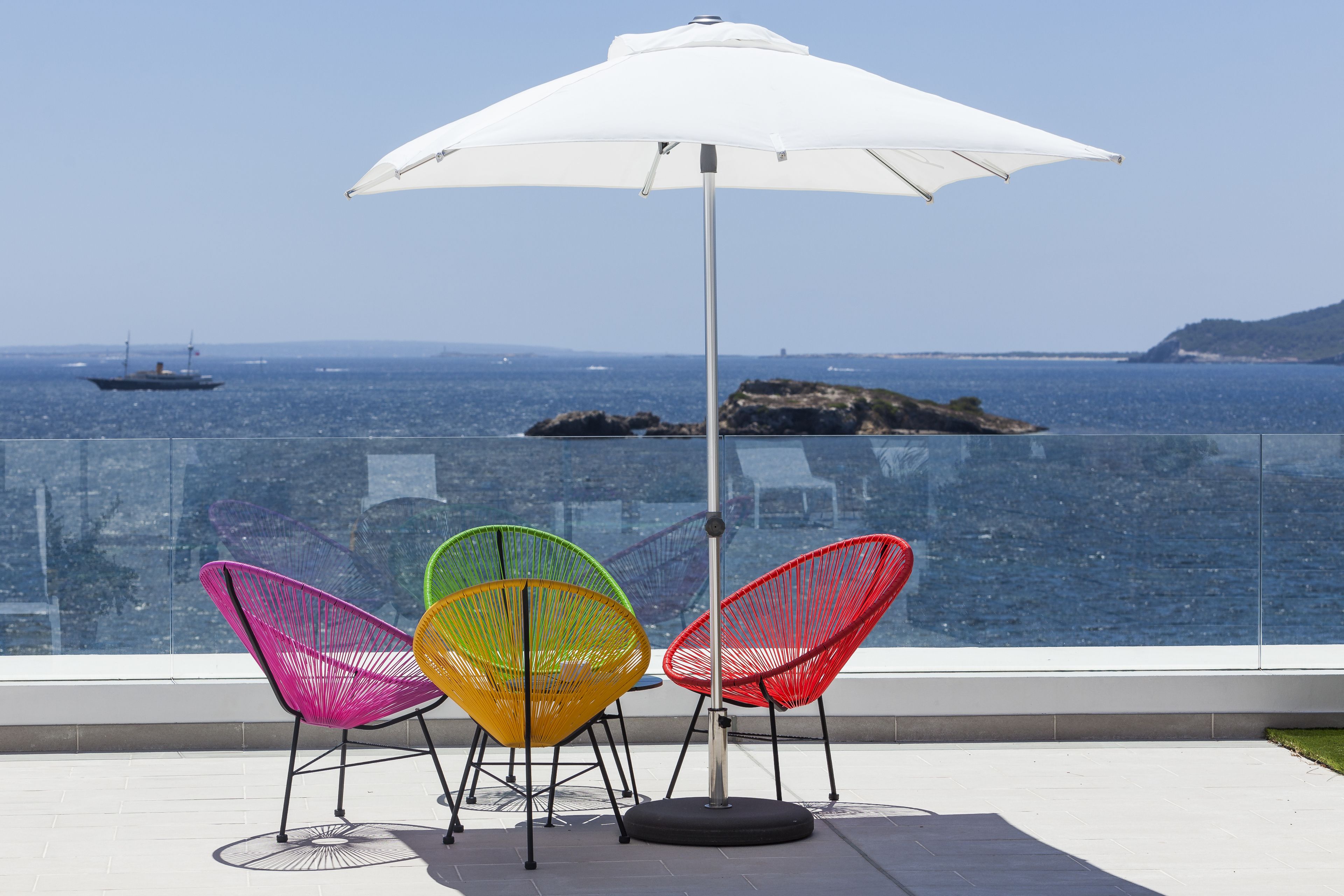 A balcony with a white umbrella, three colorful chairs, and a view of the sea.